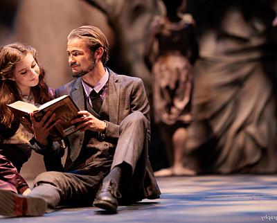 A man 和 woman sit closely in the foreground looking at a book, while two figures lurk in the background.